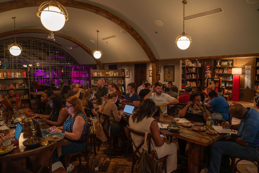 Atlanta Writes- A warmly lit library café doubles as a writing studio, with people at wooden tables eating, drinking, and working on laptops. Shelves of books line the walls, lively groups chat, and purple stage lights glow in the background.
